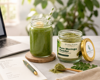 Jars of Ganga Farms Pure Moringa Powder and a green smoothie on a table with a laptop and notebook.